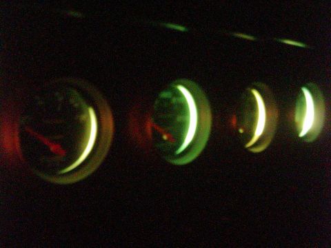 1978 Ford Bronco - Night shot of a row of four round auxiliary gauges glowing green in a dark cabin, with red needles faintly lit against the black faces. The…