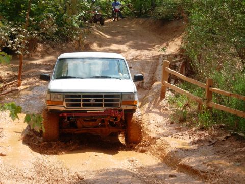 1995 Ford Bronco - A white Ford Bronco navigates a challenging muddy off-road course, with other vehicles in the distance.