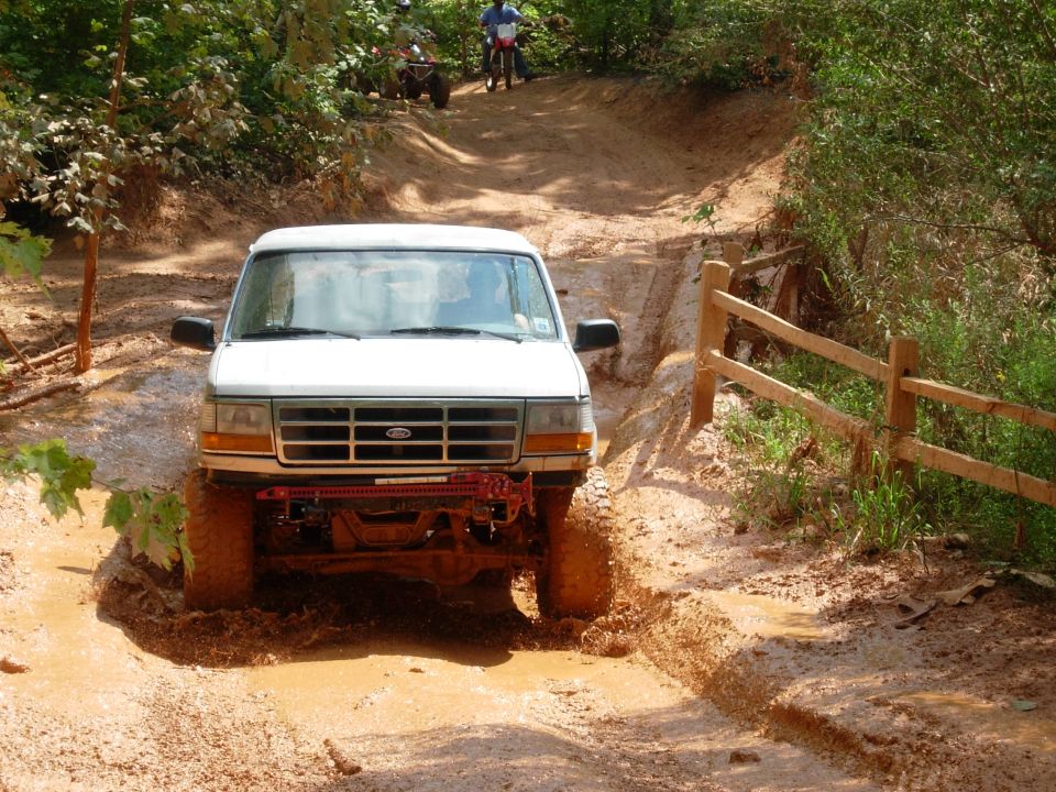 White Ford Bronco driving through a muddy trail, with an ATV and dirt bike visible in the background