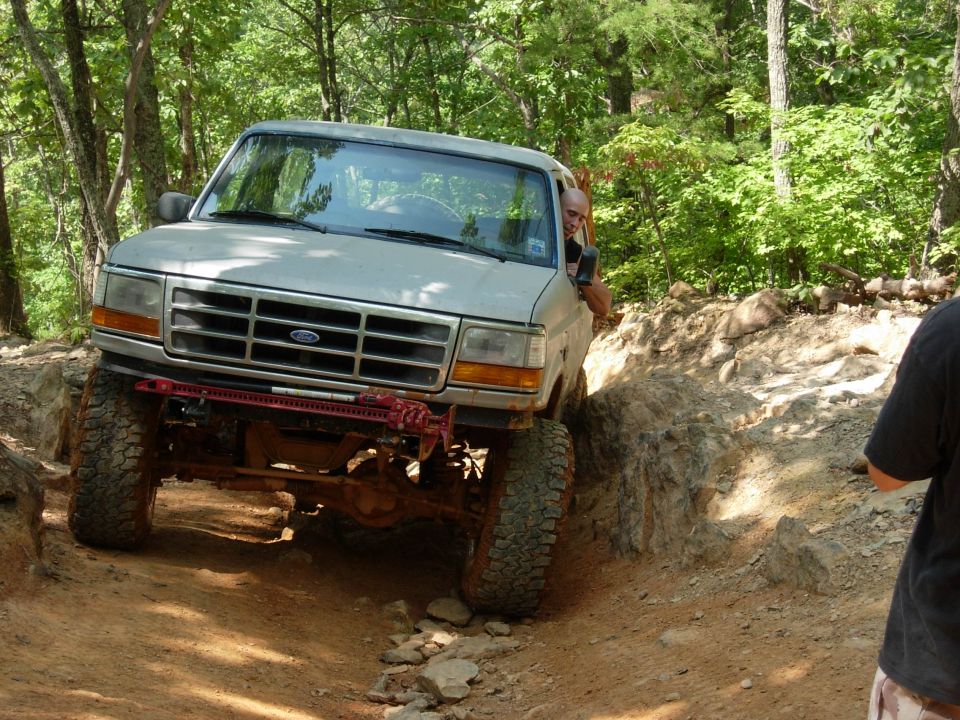 White Ford pickup climbing a rocky off-road ledge in the woods with winch mount and mud tires