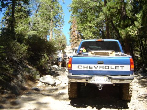 1991 Ford Bronco - Two trucks navigate a rough, wooded trail on a sunny day.