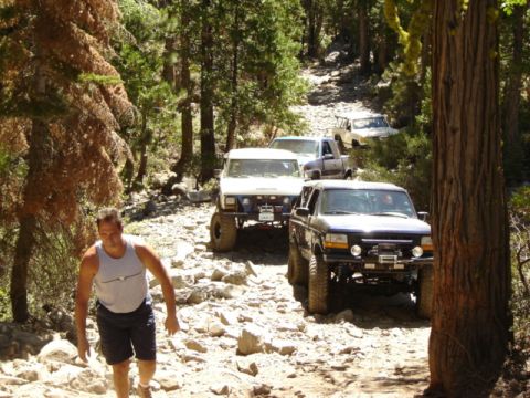 1991 Ford Bronco - A group of off-road vehicles, including a black 1991 Ford Bronco, navigate a challenging rocky path through a wooded area.