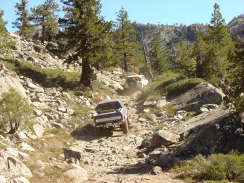 1991 Ford Bronco - Two trucks navigate a challenging off-road path through a mountainous, wooded area.