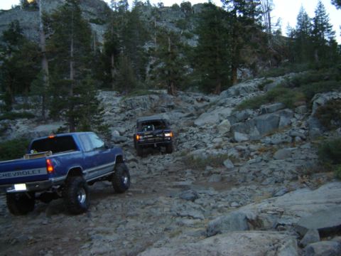 1991 Ford Bronco - Two trucks navigate a challenging, rocky mountain path surrounded by trees.