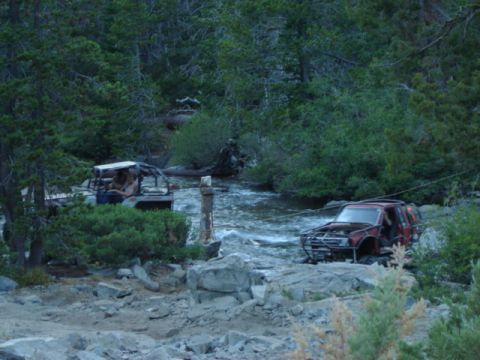 1991 Ford Bronco - Two off-road vehicles navigate a rocky stream surrounded by dense forest.