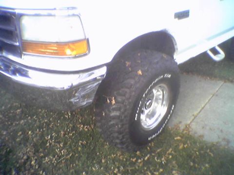 1993 Ford Bronco - Front corner of a white Ford Bronco with off-road tires and a damaged chrome bumper.