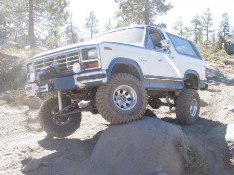 1986 Ford Bronco - White Ford Bronco with blue pinstriping and a removable hardtop climbing a granite boulder on a sandy trail, viewed from the driver 3/4 fro…