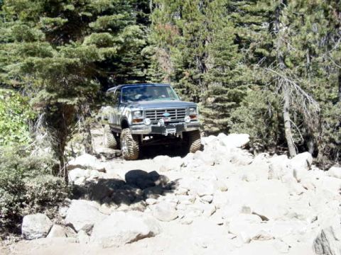 1986 Ford Bronco - Blue lifted Ford 4x4 with a white roof easing down a white granite rock trail through a pine forest, shot from a distance head-on. A winch…