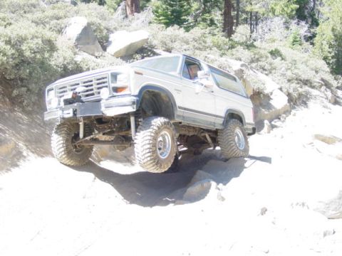 1986 Ford Bronco - Lifted white and blue Ford Bronco flexing over granite boulders on a sandy trail, seen from the driver 3/4 front with a wheel stuffed and t…