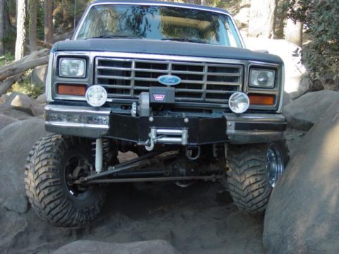 1986 Ford Bronco - Head-on view of a lifted 1980s Ford Bronco crawling between large boulders on a dirt trail, its solid front axle articulating over the rock…
