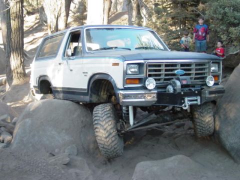 1986 Ford Bronco - A white and dark-gray two-door Ford Bronco SUV climbing over large boulders on a rocky off-road trail, shown from the front-right three-qua…