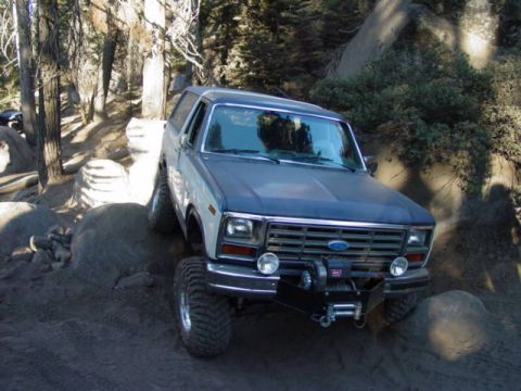 1986 Ford Bronco - High-angle 3/4 front view of a white and blue Ford Bronco full-size SUV with a removable hardtop, parked on a dirt and granite trail among…