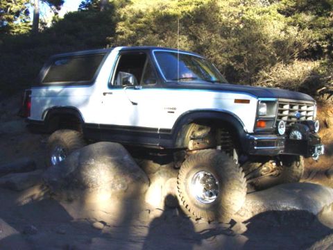 1986 Ford Bronco - White and blue two-tone Ford Bronco full-size SUV with removable hardtop crawling over granite slabs on a wooded trail, shot from the passe…