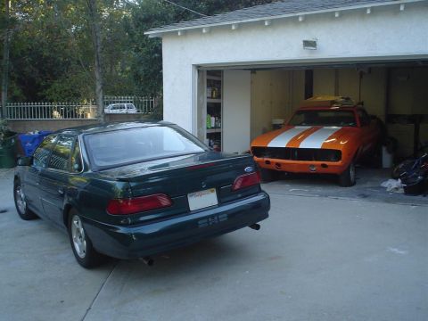 1993 Ford Taurus - A 1993 Ford Taurus is parked outside a garage, which contains an orange muscle car.