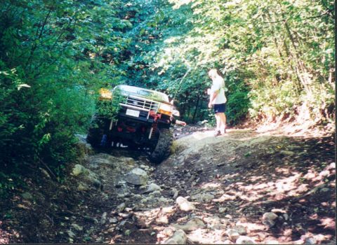 1993 Ford Bronco - A person watches as a 1993 Ford Bronco tackles a challenging, rocky off-road path surrounded by dense trees.