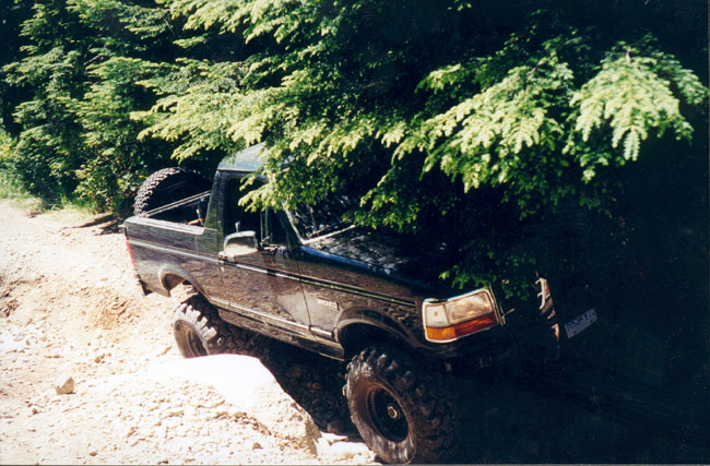 Black 1993 Ford Bronco with large tires driving off-road through trees