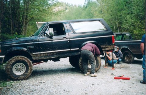 1993 Ford Bronco - A group of people work on the rear tire of a black 1993 Ford Bronco, with another truck visible in the background.