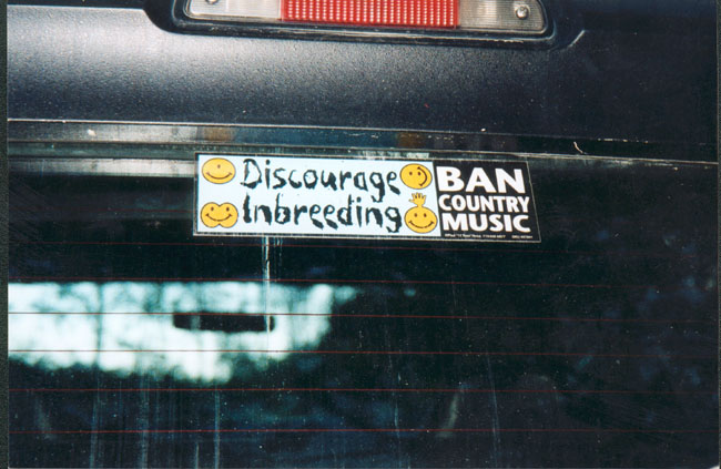 Rear window of a dark vehicle with two bumper stickers: one says 'Discourage Inbreeding' with smiley faces, the other says 'BAN COUNTRY MUSIC' with smiley faces