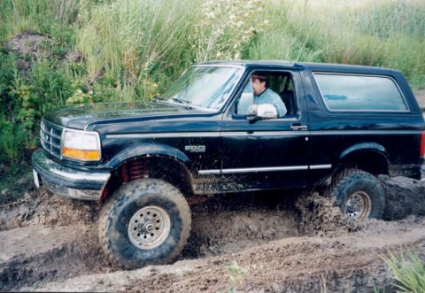1993 Ford Bronco - A black 1993 Ford Bronco navigates a muddy off-road trail, kicking up dirt as it goes.