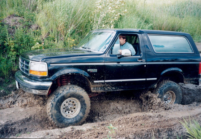 Black 1993 Ford Bronco with lifted suspension driving through mud, driver visible inside