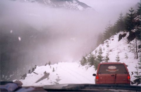 1993 Ford Bronco - A red SUV navigates a snow-covered mountain road, with fog obscuring the distant peaks.