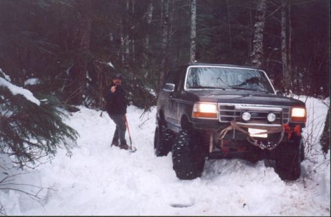 1993 Ford Bronco - A lifted Ford Bronco navigates a snowy forest trail with a person in snowshoes observing.