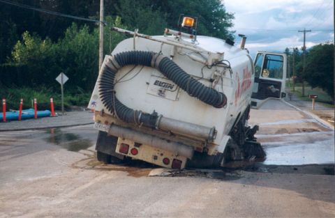 1993 Ford Bronco - A street sweeper truck is shown with its large vacuum hose and brushes deployed, possibly stuck on uneven terrain.