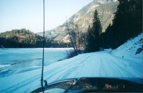 1993 Ford Bronco - View from inside a vehicle on a snowy road beside a frozen lake, with mountains visible in the distance.