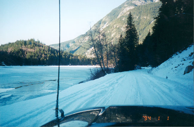 Dashboard view from a vehicle driving on a snowy road beside a frozen lake, with mountains in the background