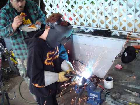 1993 Ford Bronco - A person welds with sparks flying, while another person stands nearby eating pizza.