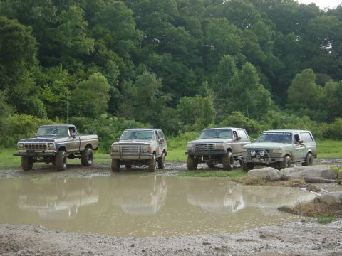 1989 Ford Bronco - Several off-road vehicles are lined up in a muddy clearing, with trees in the background.