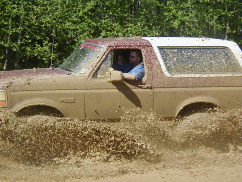 Mud-covered Ford Bronco blasting through a mud pit, two occupants visible, mud spraying