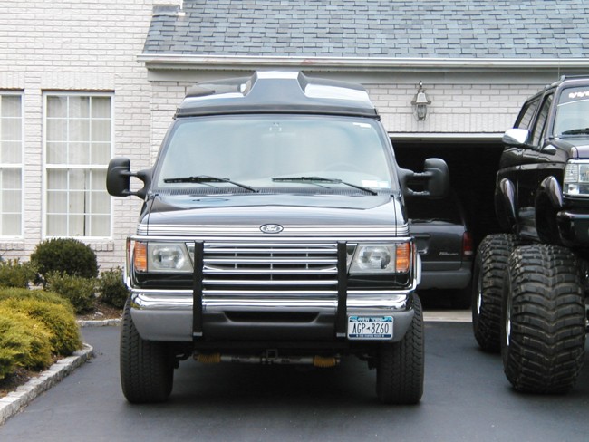 Front of dark gray Ford high-top van with grille guard and NY plate in driveway