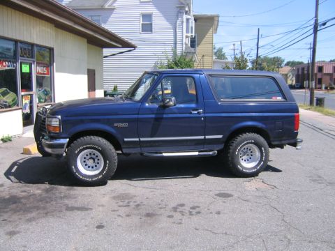 1994 Ford Bronco - A blue Ford Bronco SUV is parked on a cracked asphalt lot in front of a building.