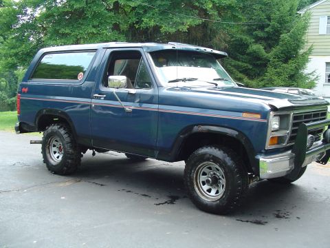 1986 Ford Bronco - This blue Ford Bronco is parked on a paved surface with trees and a house in the background.