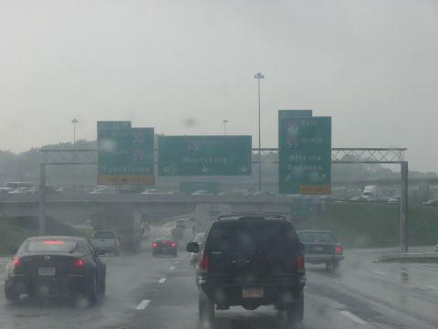 1988 Ford Bronco - Traffic moves on a wet highway under overhead signs pointing to various cities.