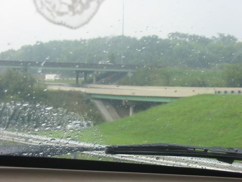1988 Ford Bronco - Raindrops on the windshield obscure the view of highway overpasses and green hills.