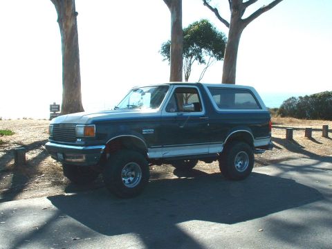 1990 Ford Bronco - A lifted 1990 Ford Bronco is seen from a three-quarter front angle, parked outdoors with trees and a coastal view in the background.