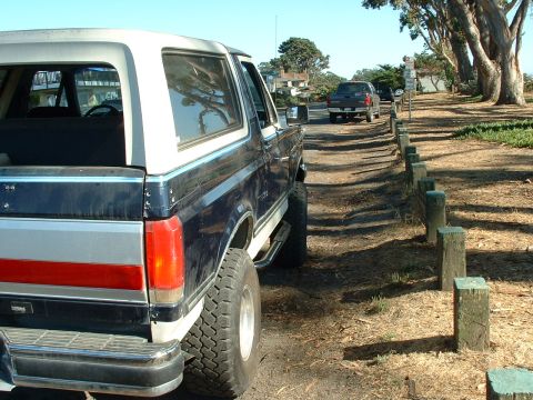 1990 Ford Bronco - A lifted Ford Bronco is parked on a dirt road next to a line of wooden posts, with another truck in the distance.