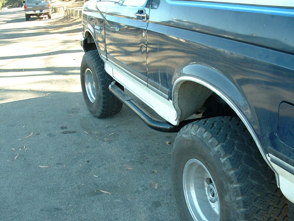 1990 Ford Bronco side view with running boards and large tires. Another truck is visible in the background