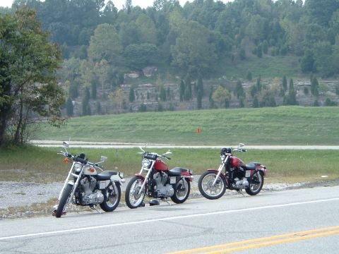 1995 Mercury Grand Marquis - Three motorcycles are parked on the side of a road with a grassy hillside behind them.