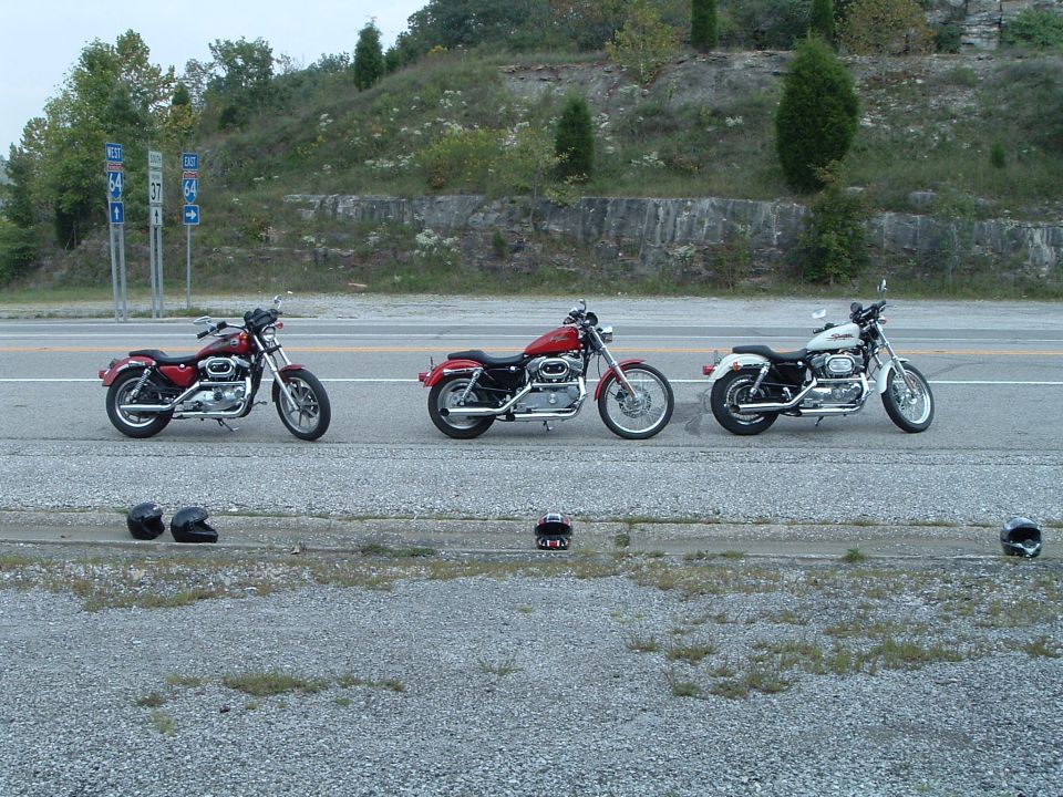 Three Harley Sportsters lined up on a highway shoulder with helmets on the gravel