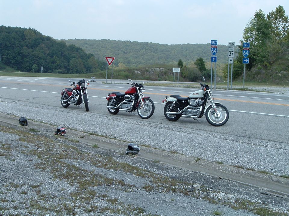 Three Harley Sportster motorcycles parked along a wooded highway near Route 64 signs