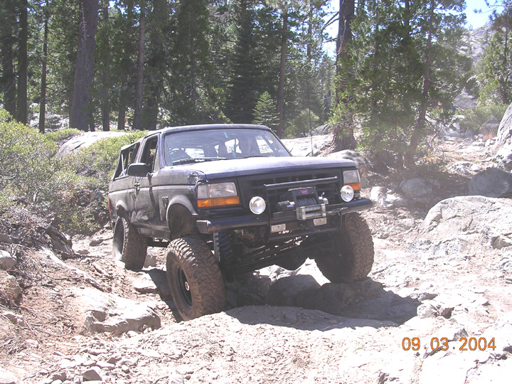Dark blue topless Ford Bronco with winch bumper crawling granite rocks on a pine forest trail