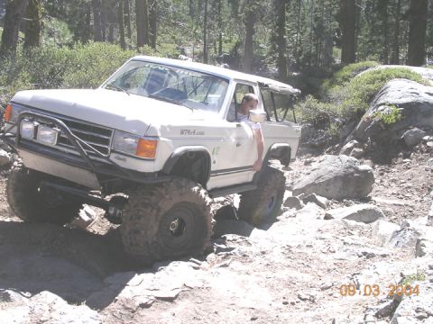 1994 Ford Bronco - A modified off-road vehicle tackles a challenging rocky path in a wooded area.