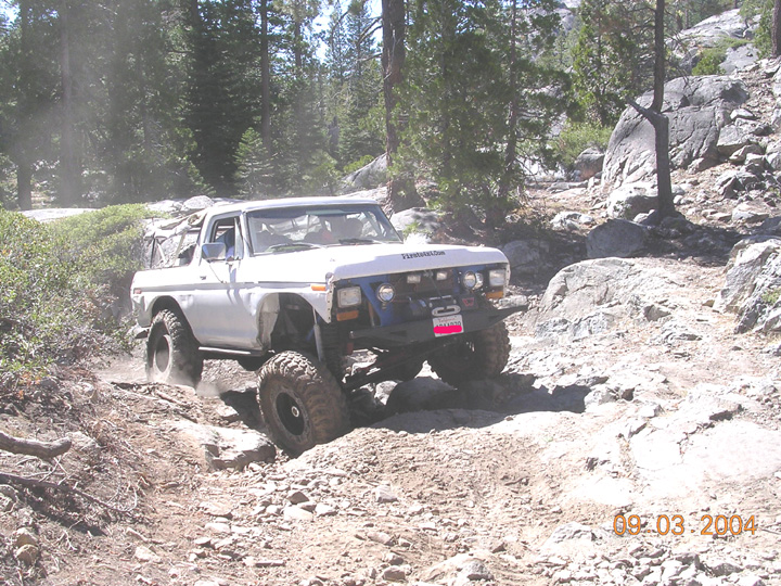 White off-road SUV with large tires navigating a rocky trail. It has a winch on the front bumper and a blue grille insert