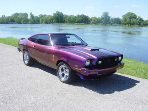 1978 Ford Mustang II - This 1978 Ford Mustang II coupe, painted in a striking purple hue, features a prominent hood scoop and is captured in a three-quarter front…
