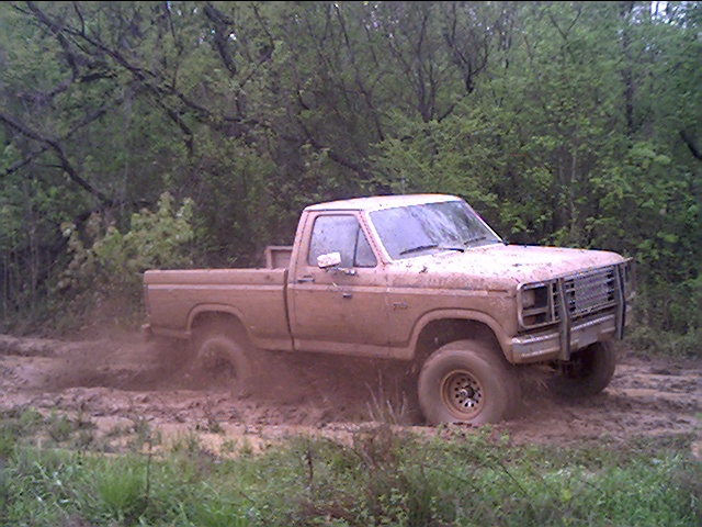 Mud-covered early-1980s Ford pickup spinning through a wooded mud hole with brush guard