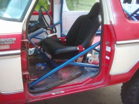 1978 Ford Bronco - Open doorway of a red and white Bronco Ranger XLT mid-build, showing a blue tube roll cage running through the cab and a black high-back bu…