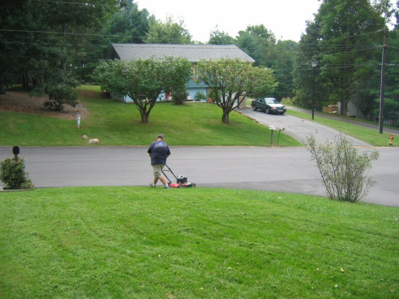 Man mowing a green lawn in front of a house with a car parked in the driveway